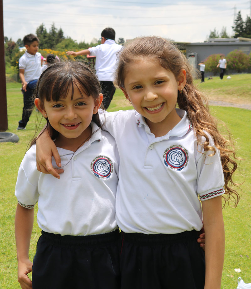 Dos niñas sonrientes con uniforme escolar blanco y pantalones oscuros, abrazadas en un campo verde con otros niños jugando al fondo.