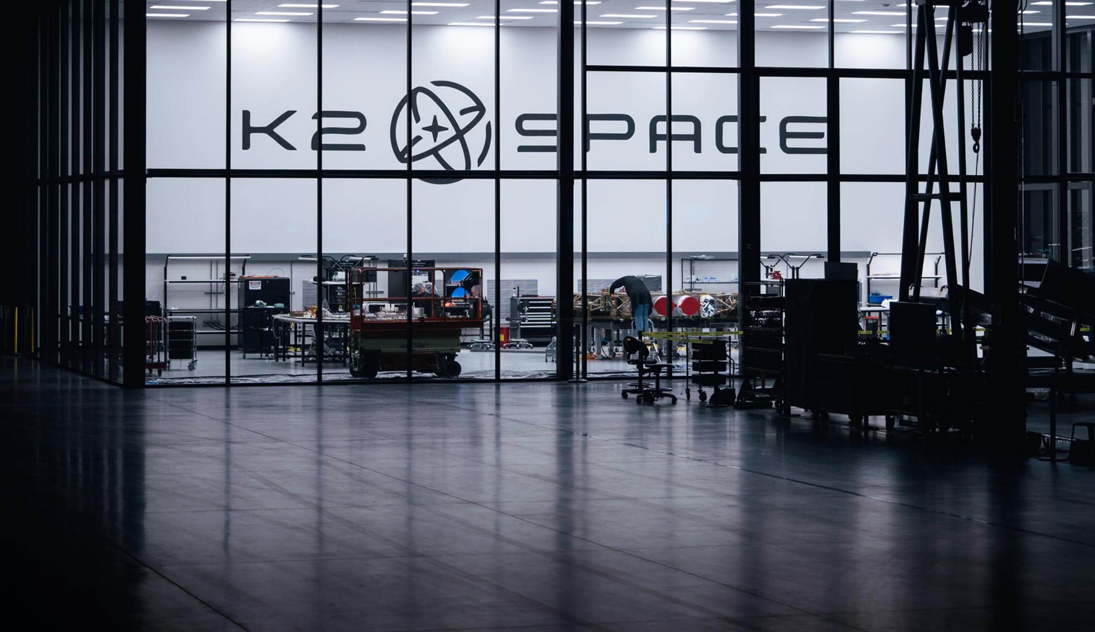 Engineers working inside the K2 Space manufacturing facility, assembling satellite components behind large glass panels