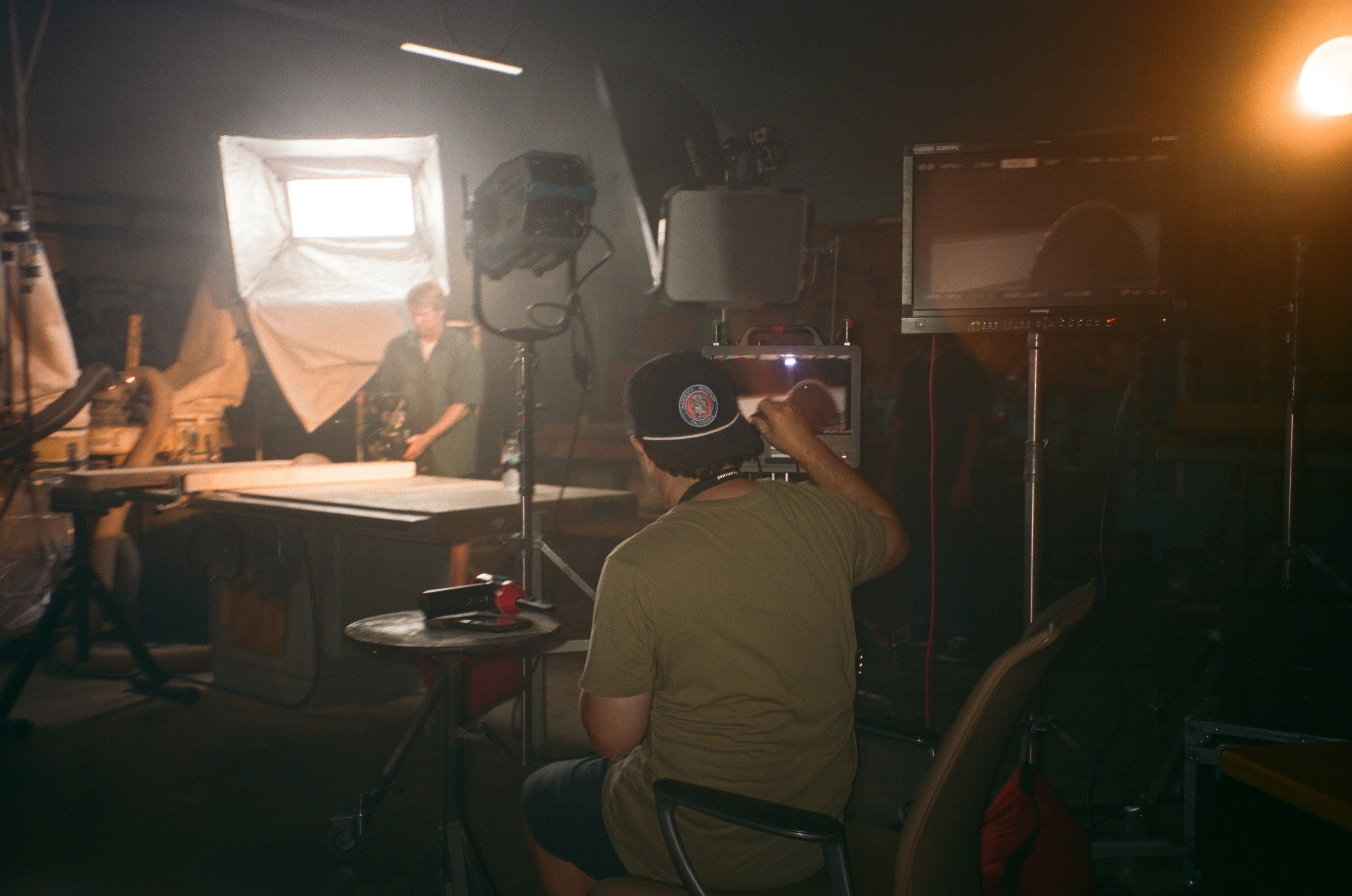 Man sitting in a chair monitoring a video screen on a film set with lighting equipment and another person working in the background.