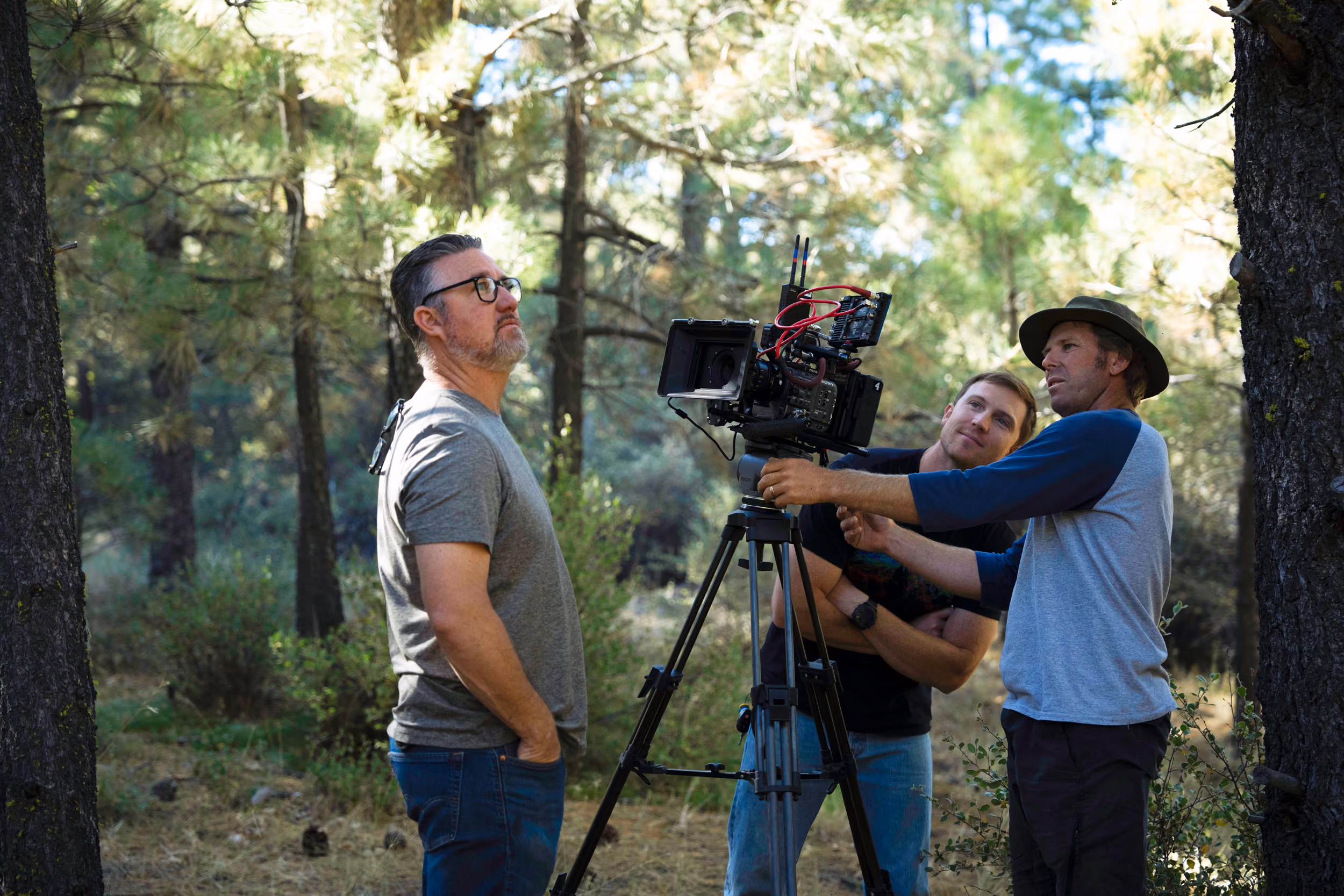 Three men outdoors in a forest setting, one operating a professional video camera on a tripod, one observing, and one posing for the camera.