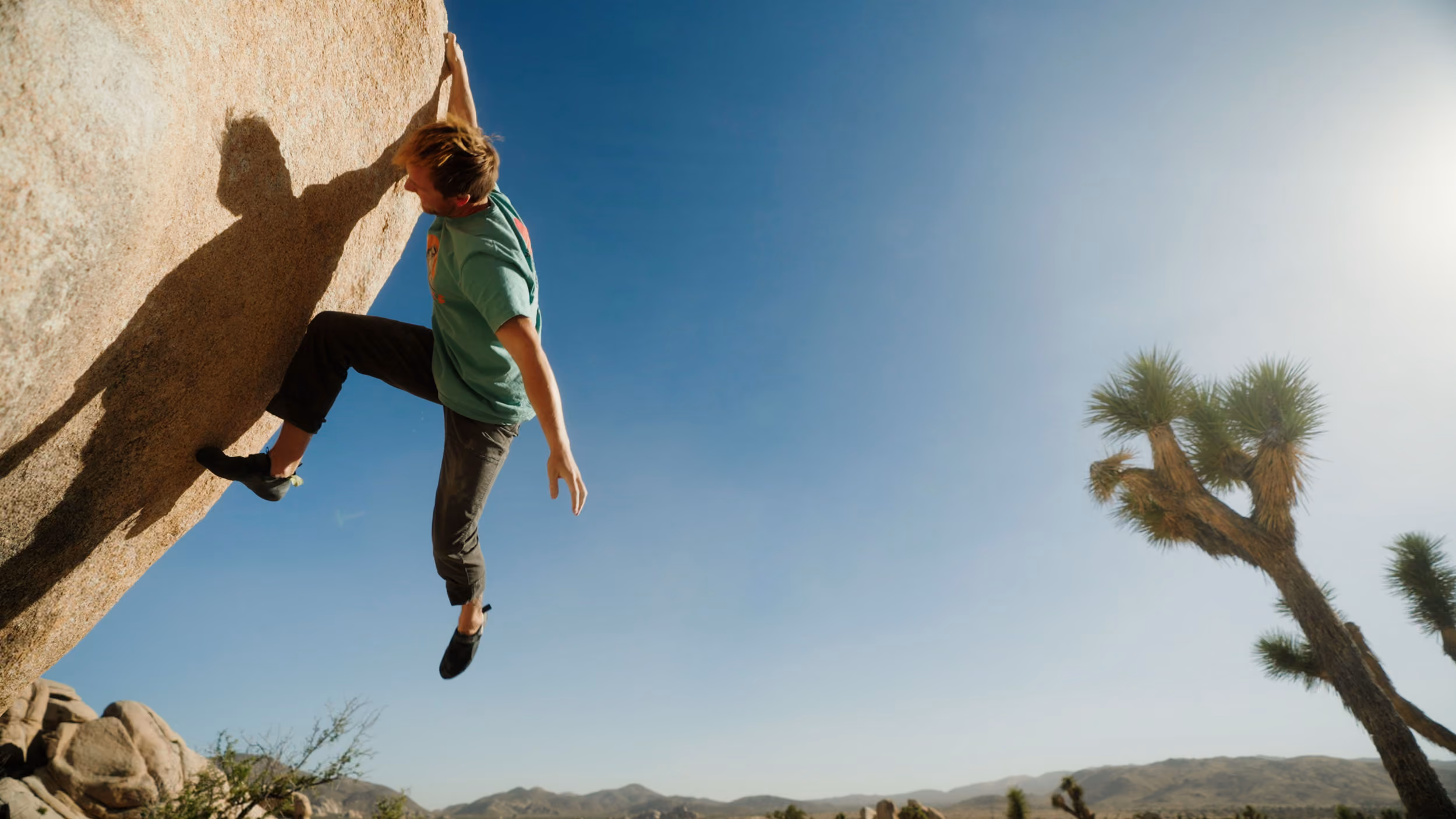 Man climbing a large rock in a desert landscape with a clear blue sky and Joshua trees.