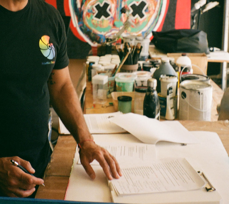 Person wearing black shirt writing on papers at a cluttered table with art supplies and paint containers.