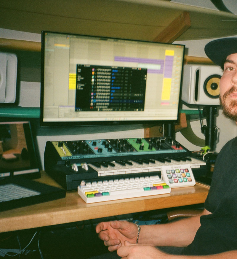 Person wearing a black hat seated at a desk with a music production setup including a keyboard, mixer, and monitor displaying audio software.