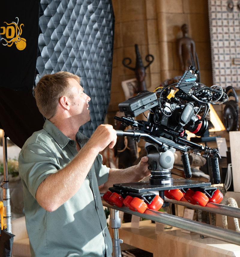 Man operating a professional video camera mounted on a slider dolly with red wheels inside a studio setting.