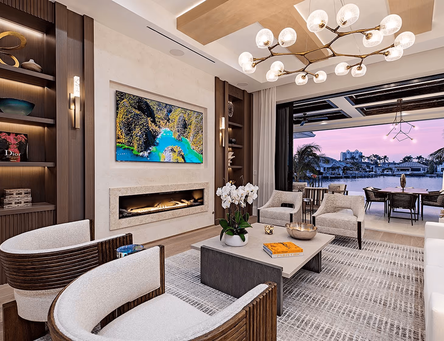 Modern living room with light wood and beige decor, a fireplace, wall-mounted TV, and large sliding doors opening to a waterfront patio at sunset.