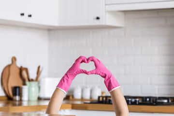 Hands wearing pink rubber gloves forming a heart shape in a modern kitchen.
