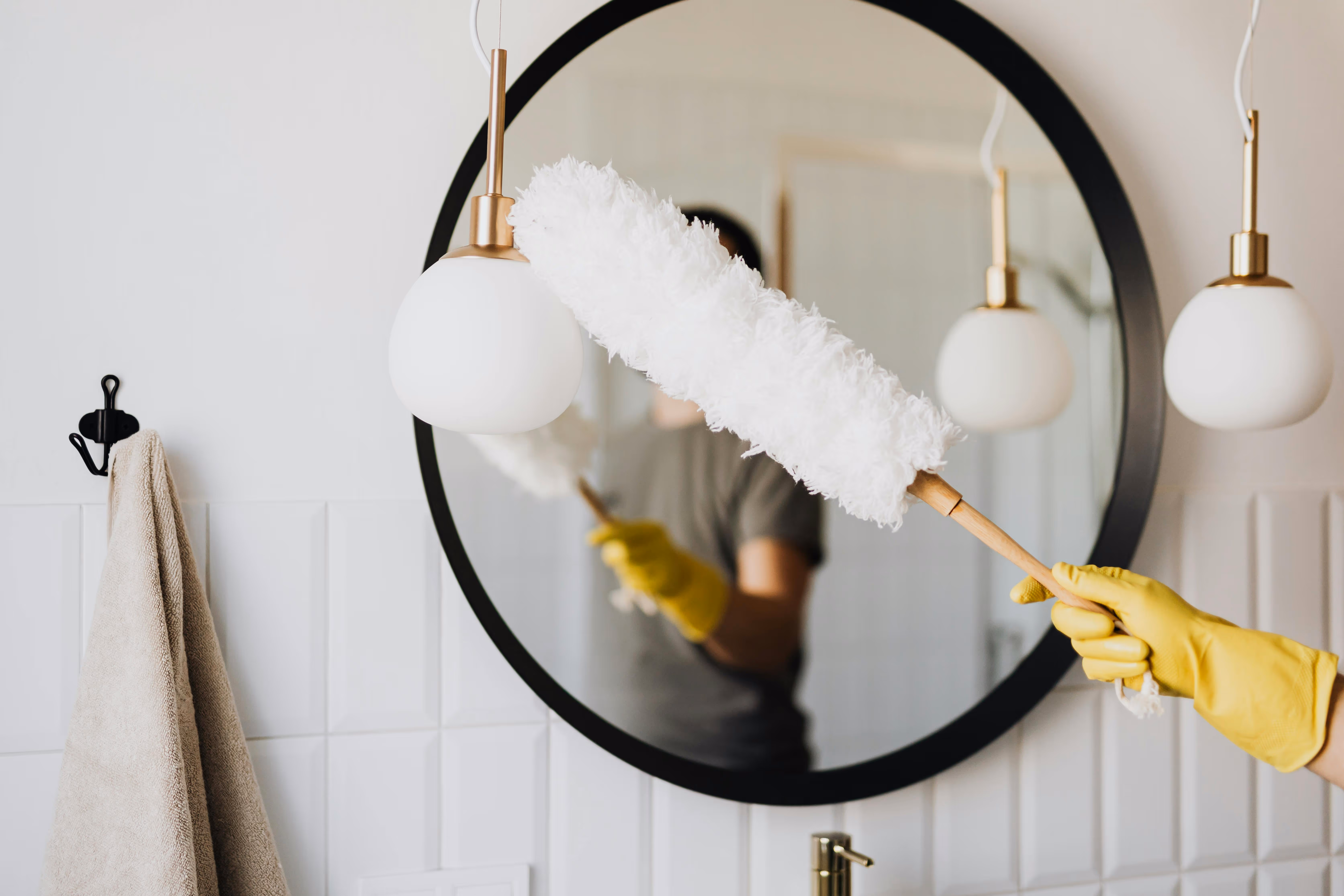 Person wearing yellow gloves dusting a round mirror in a bathroom with a white fluffy duster.