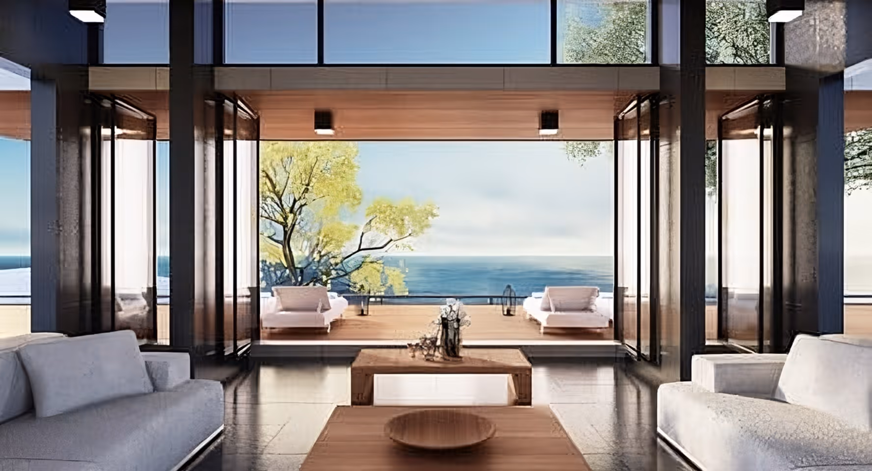 Modern living room with gray sofas facing a wooden coffee table and large open windows overlooking a deck with lounge chairs, trees, and ocean view.
