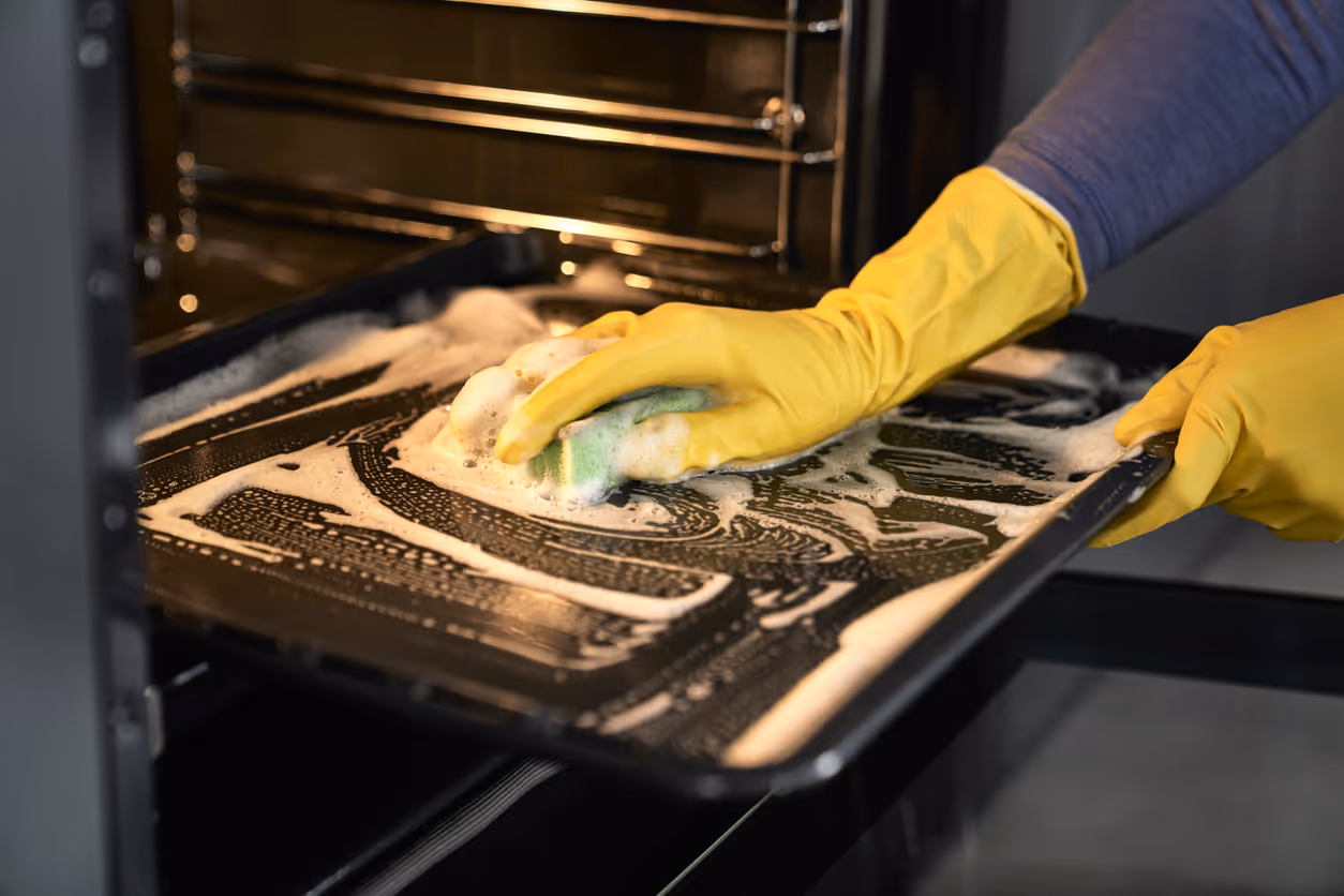 Person wearing yellow gloves scrubbing an oven tray with soapy foam inside an oven.