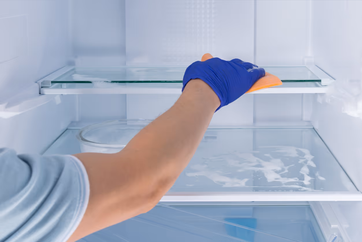 Person wearing a blue glove cleaning a glass shelf inside a refrigerator with an orange cloth.