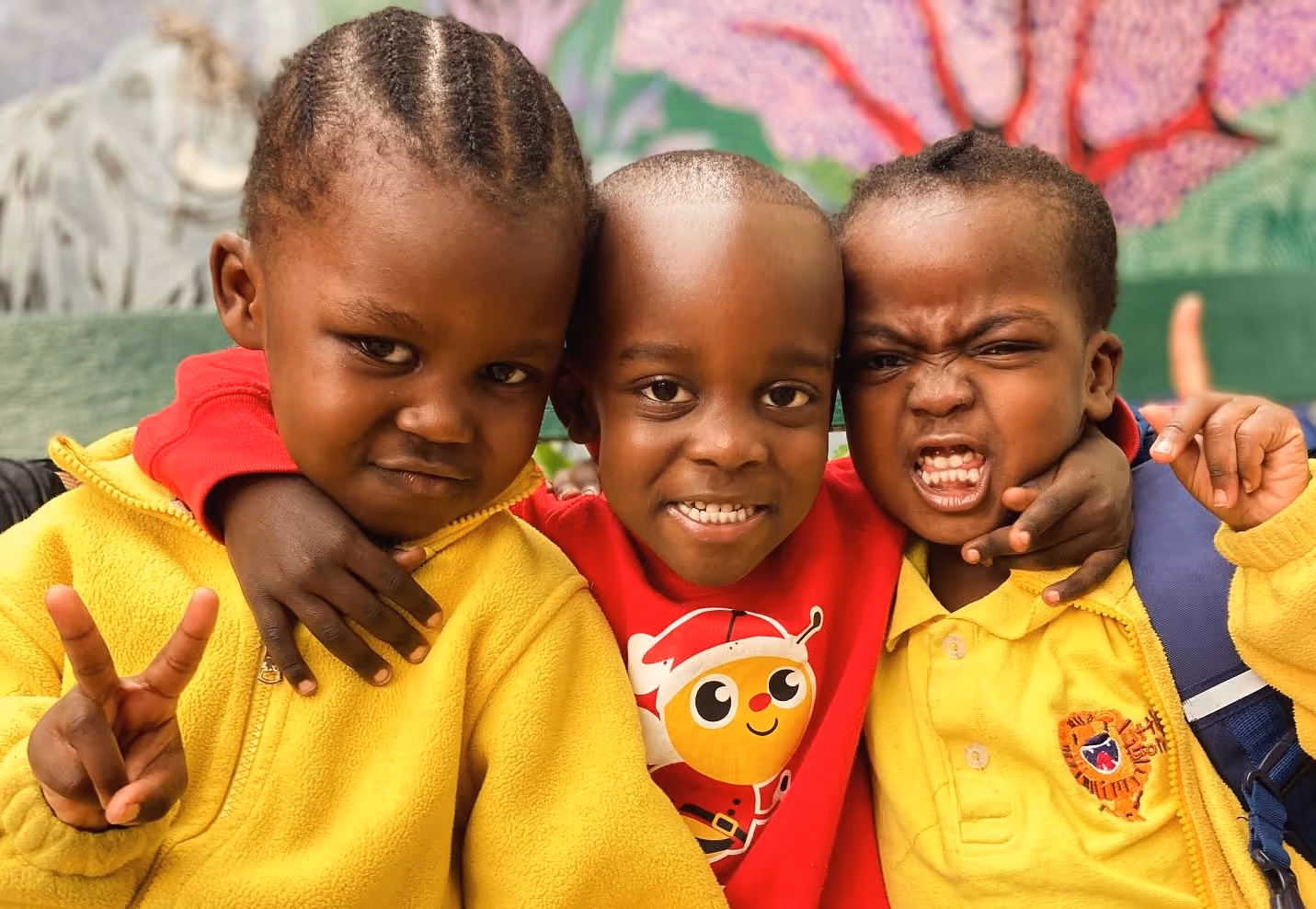 Three children smiling and posing with arms around each other, two wearing yellow jackets.