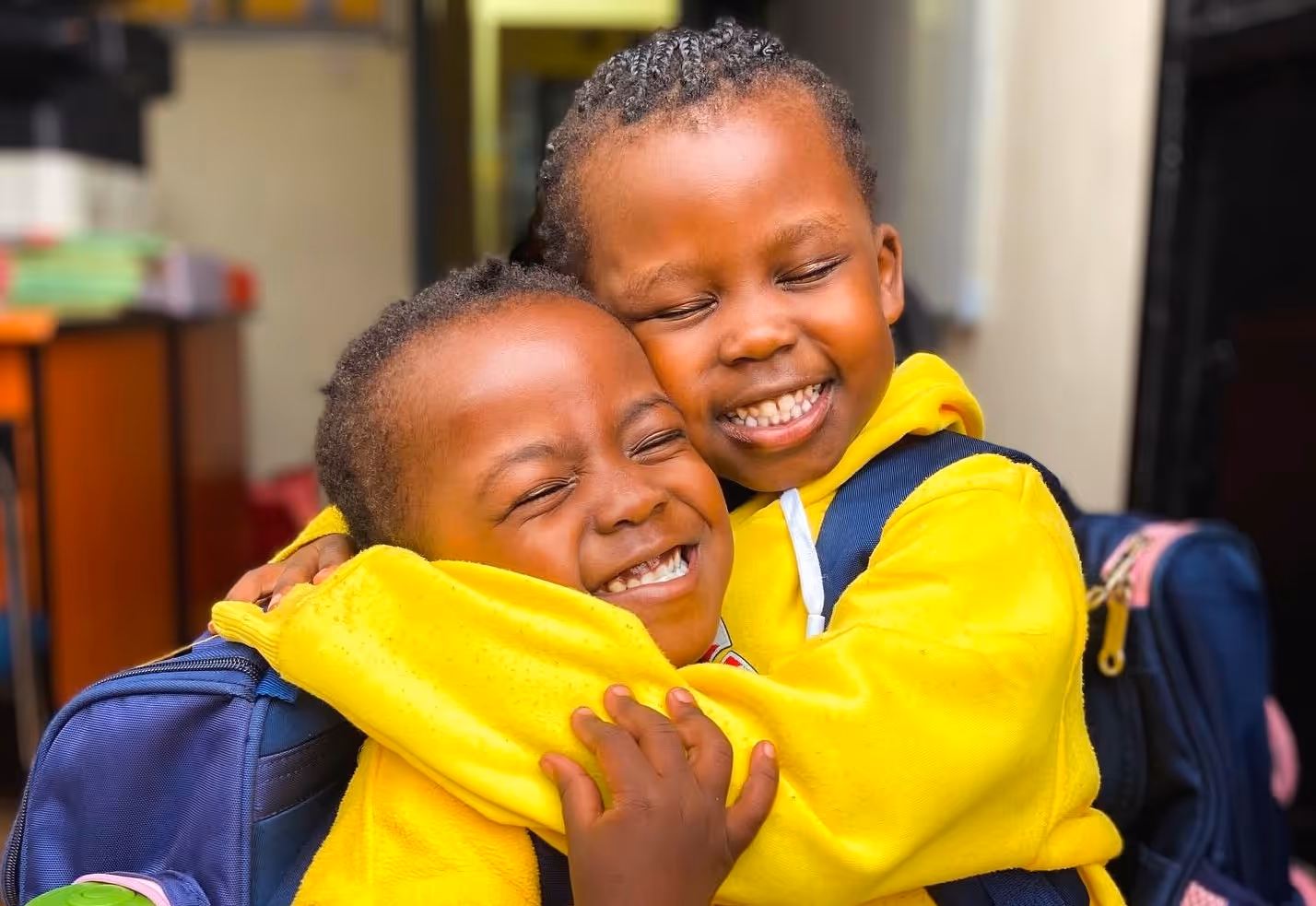 Two young children in yellow hoodies sharing a joyful hug with eyes closed and big smiles.