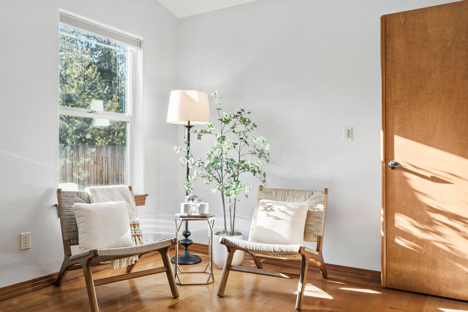 Bright corner of a room with two woven wooden chairs with white cushions and a small table with mugs between them, next to a floor lamp and a potted plant.