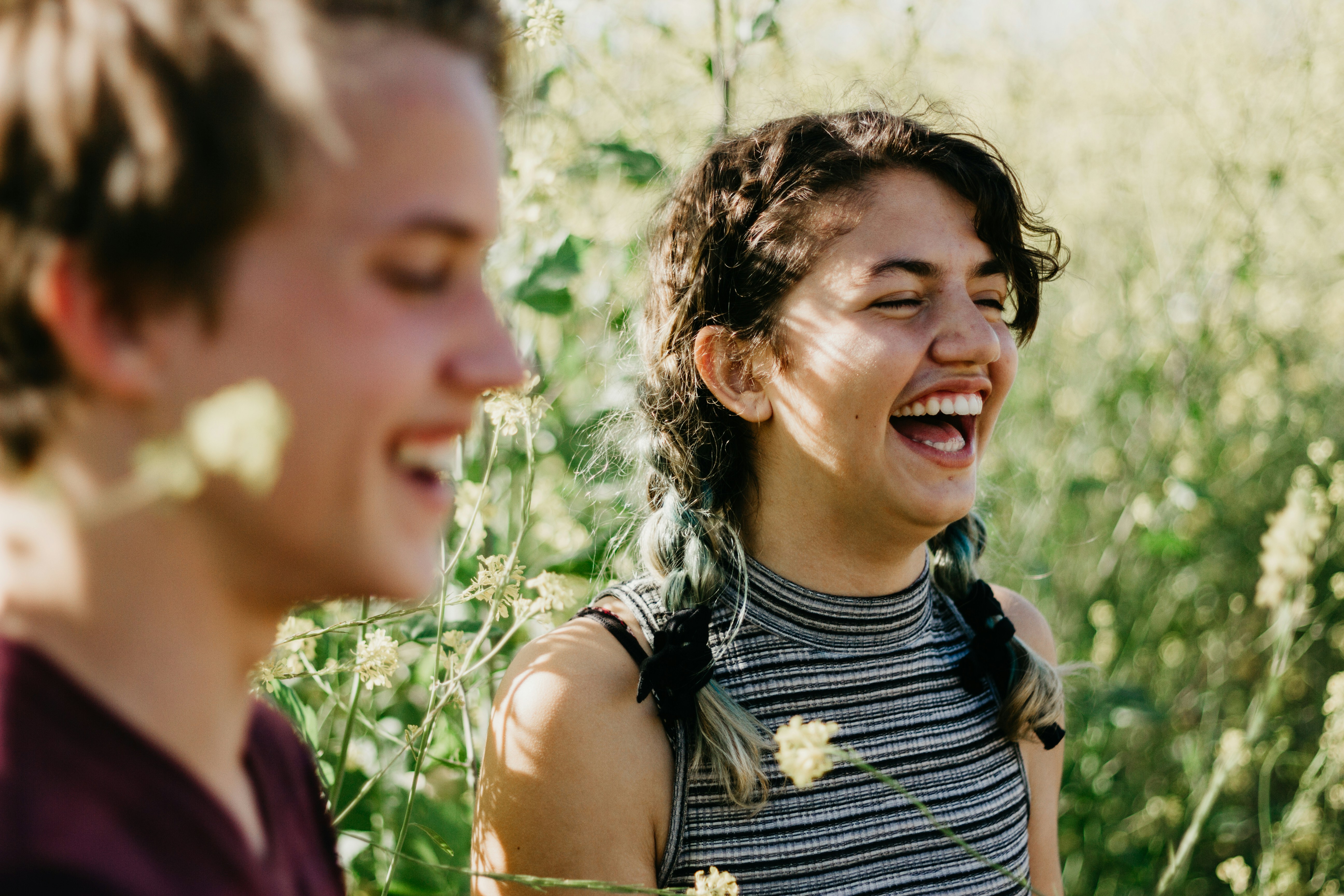 Two young people laughing outdoors in a field of wildflowers on a sunny day.
