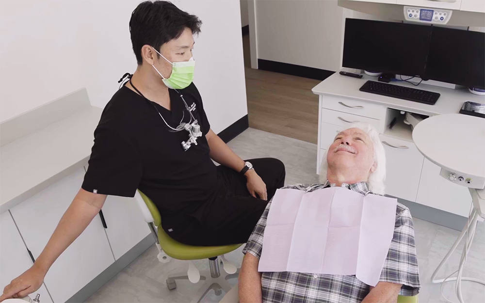 Dentist in black scrubs and green mask sitting by an elderly male patient reclining in a dental chair.