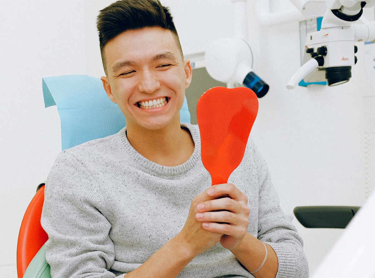 Smiling young man sitting in a dental chair holding a red hand mirror, after teeth whitening.