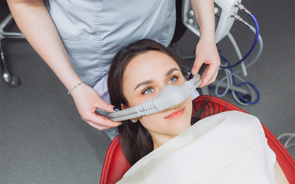 Young woman reclining in a dental chair with a nasal mask being adjusted by dental assistant.