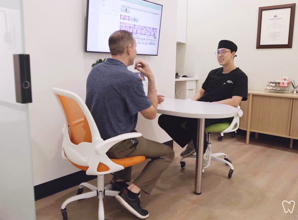 A doctor and patient sitting across from each other at a small white round table in a consultation room with a screen displaying dental images.