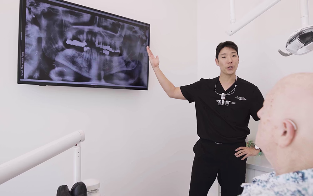 Dentist explaining dental X-ray displayed on a wall-mounted screen to a patient in a dental office.