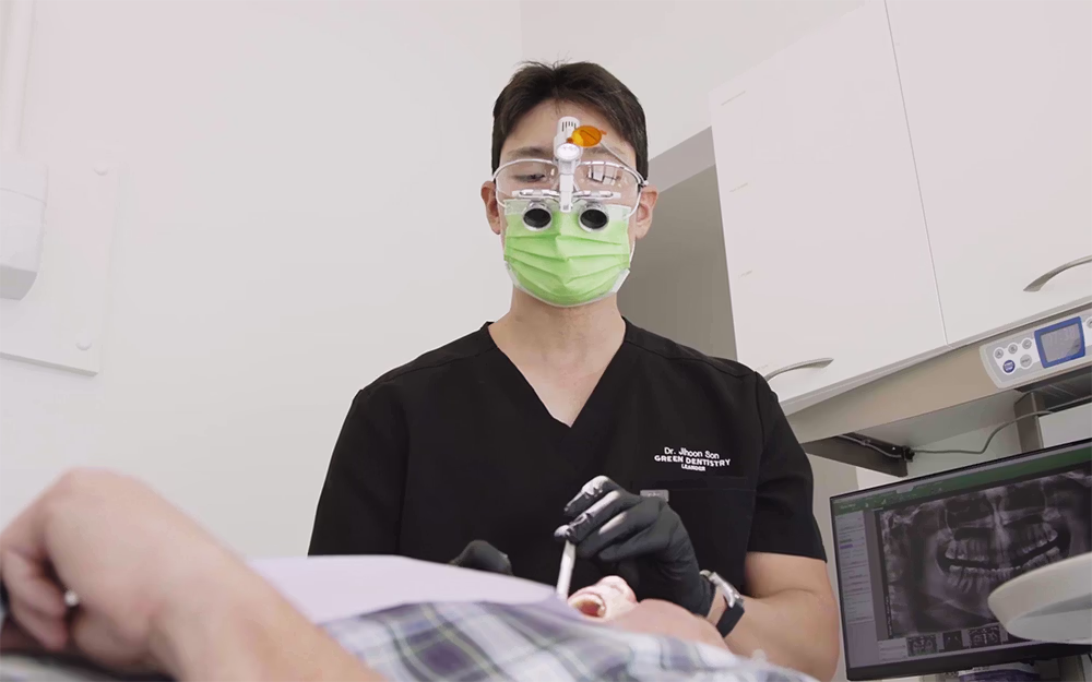 Dentist wearing magnifying glasses and green face mask treating a patient in a dental clinic with dental X-rays on a monitor.