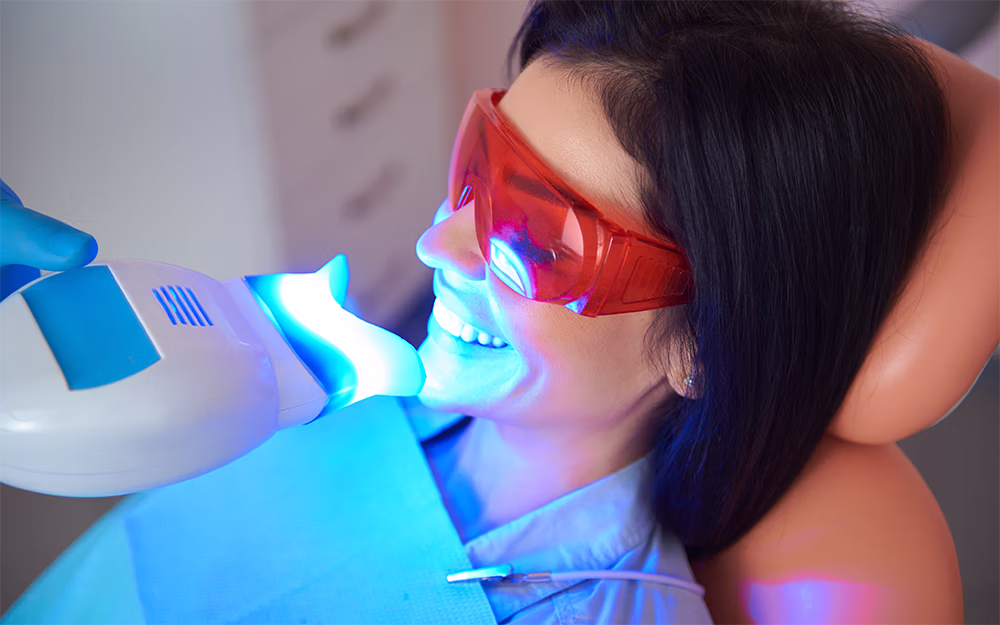 Woman wearing protective red glasses receiving dental teeth whitening treatment with a blue light device.