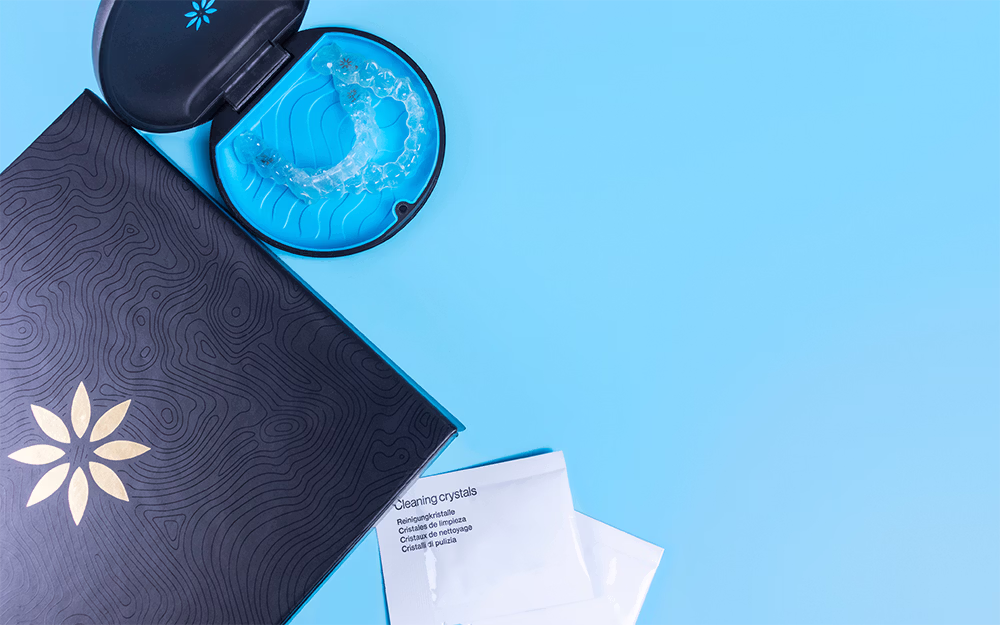 Clear dental aligners in a round black case next to a black box and white cleaning crystal packets on a blue background.