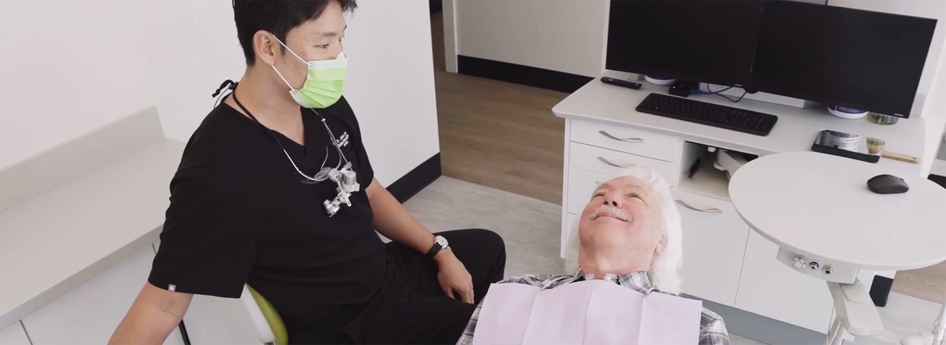 Dentist wearing a green mask interacting with an elderly patient reclined in a dental chair.