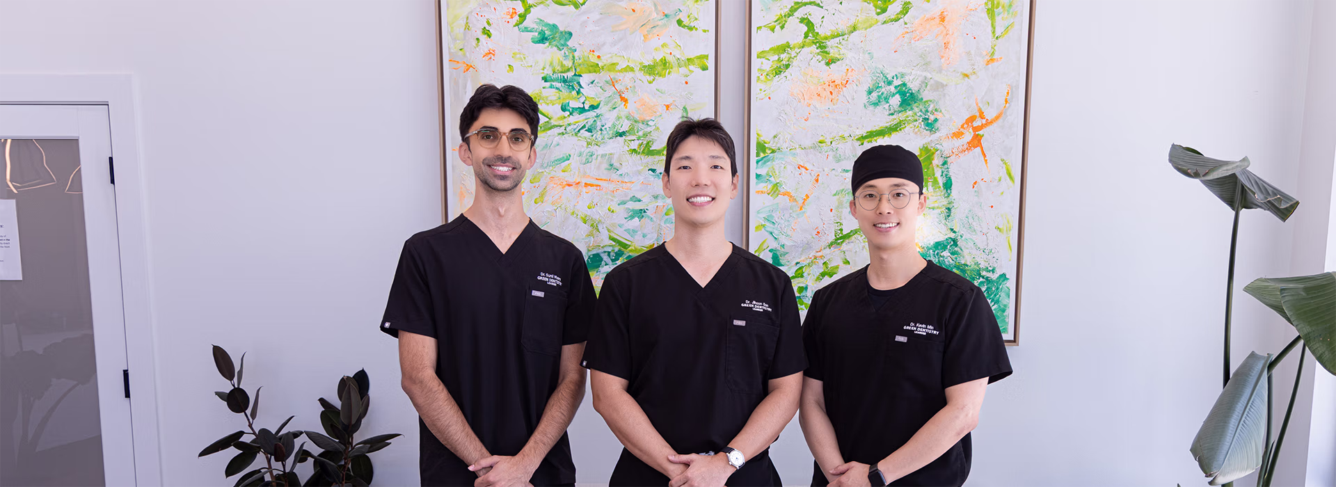 Three male dentists in black scrubs standing and smiling in a modern office with abstract paintings and plants.