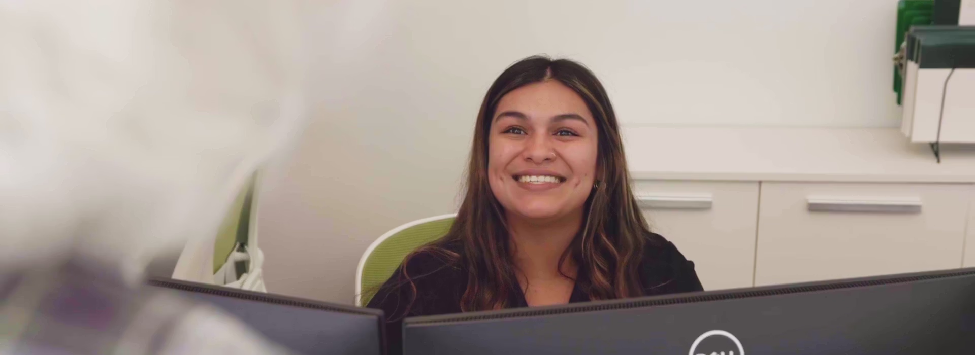 Smiling woman with long hair sitting behind computer monitors at dental reception desk.