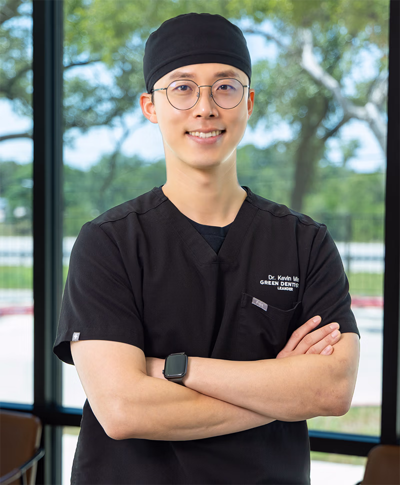 Smiling male dentist wearing black scrubs, cap, glasses, and a smartwatch standing with arms crossed inside a bright dental office.