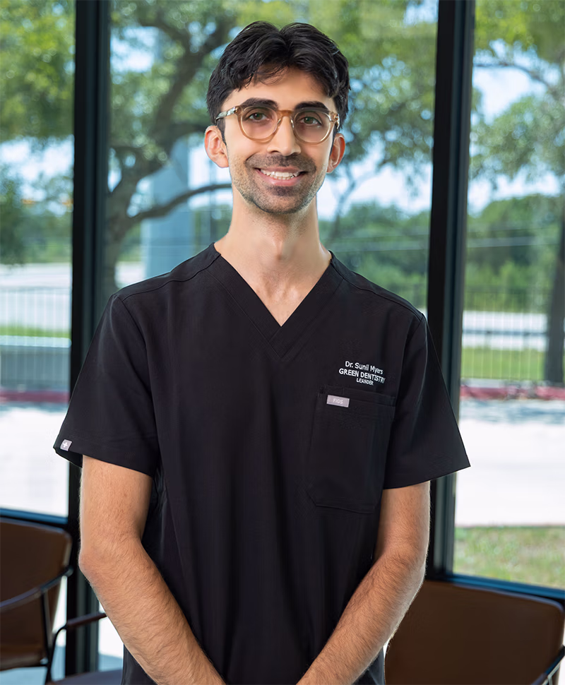 Smiling man wearing glasses and black dental scrubs standing indoors with windows and trees in the background.