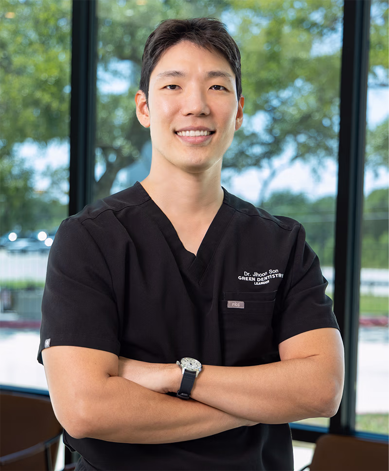 Smiling male dentist in black scrubs with arms crossed standing indoors near large windows with greenery outside.