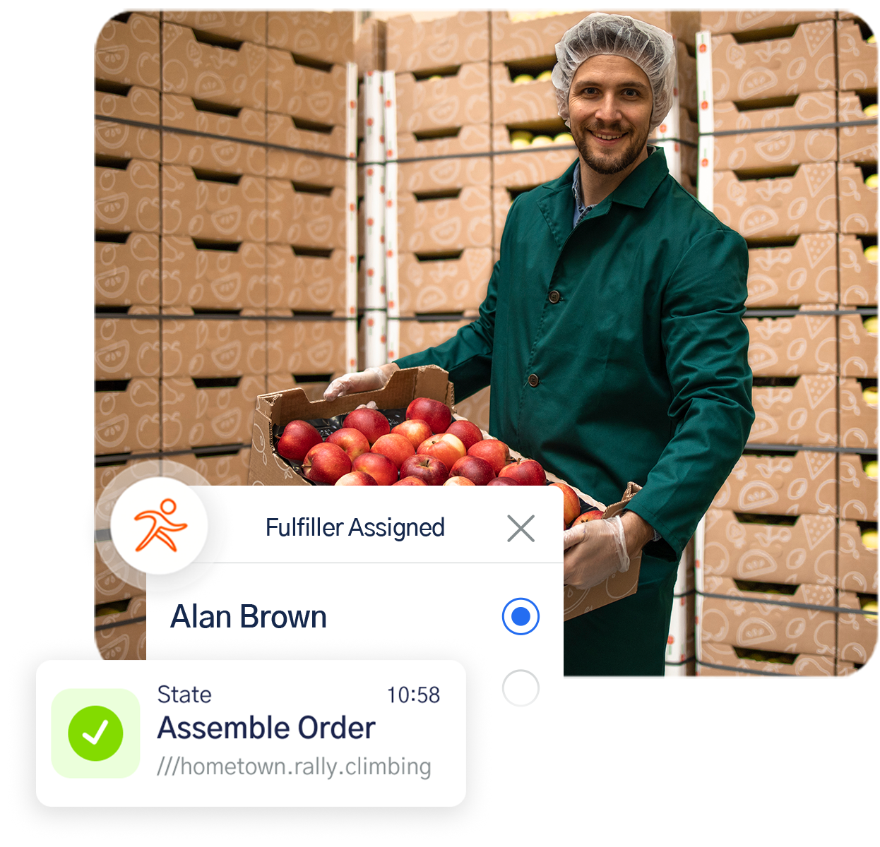 Smiling worker in green coat and hairnet holding a box of red apples in a warehouse filled with stacked fruit boxes.