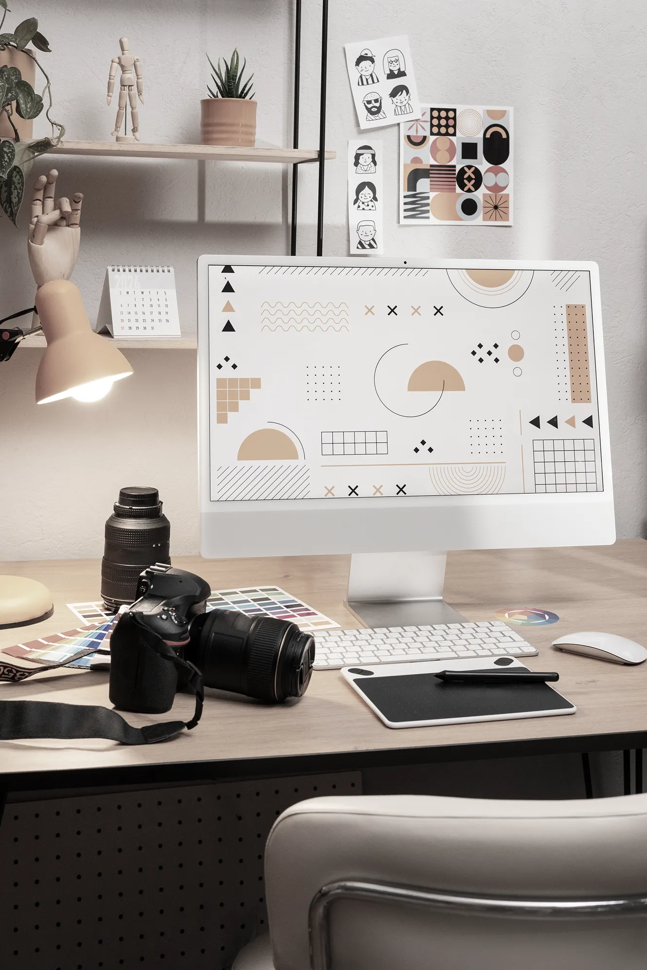Creative workspace with a computer displaying abstract geometric designs, surrounded by a camera, camera lens, drawing tablet, color swatches, and desk lamp, with shelves holding plants, figurines, and design prints on the wall.