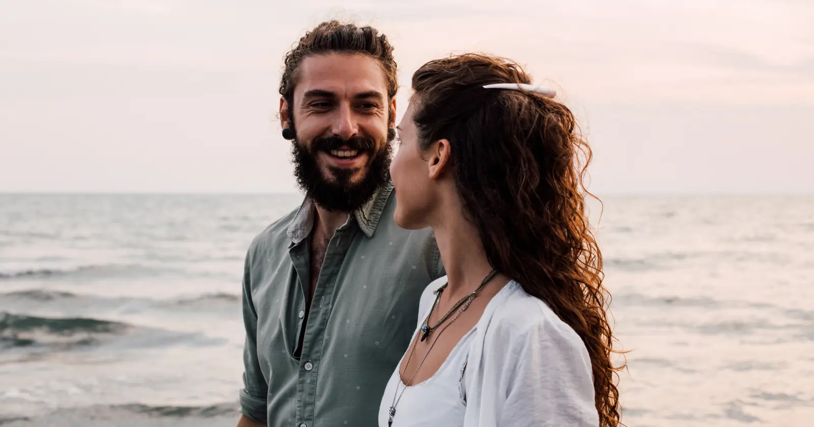 Smiling man and woman looking at each other by the ocean during sunset.