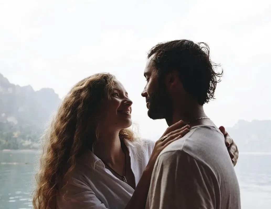 Couple embracing and smiling at each other by a calm lake with mountains in the background.