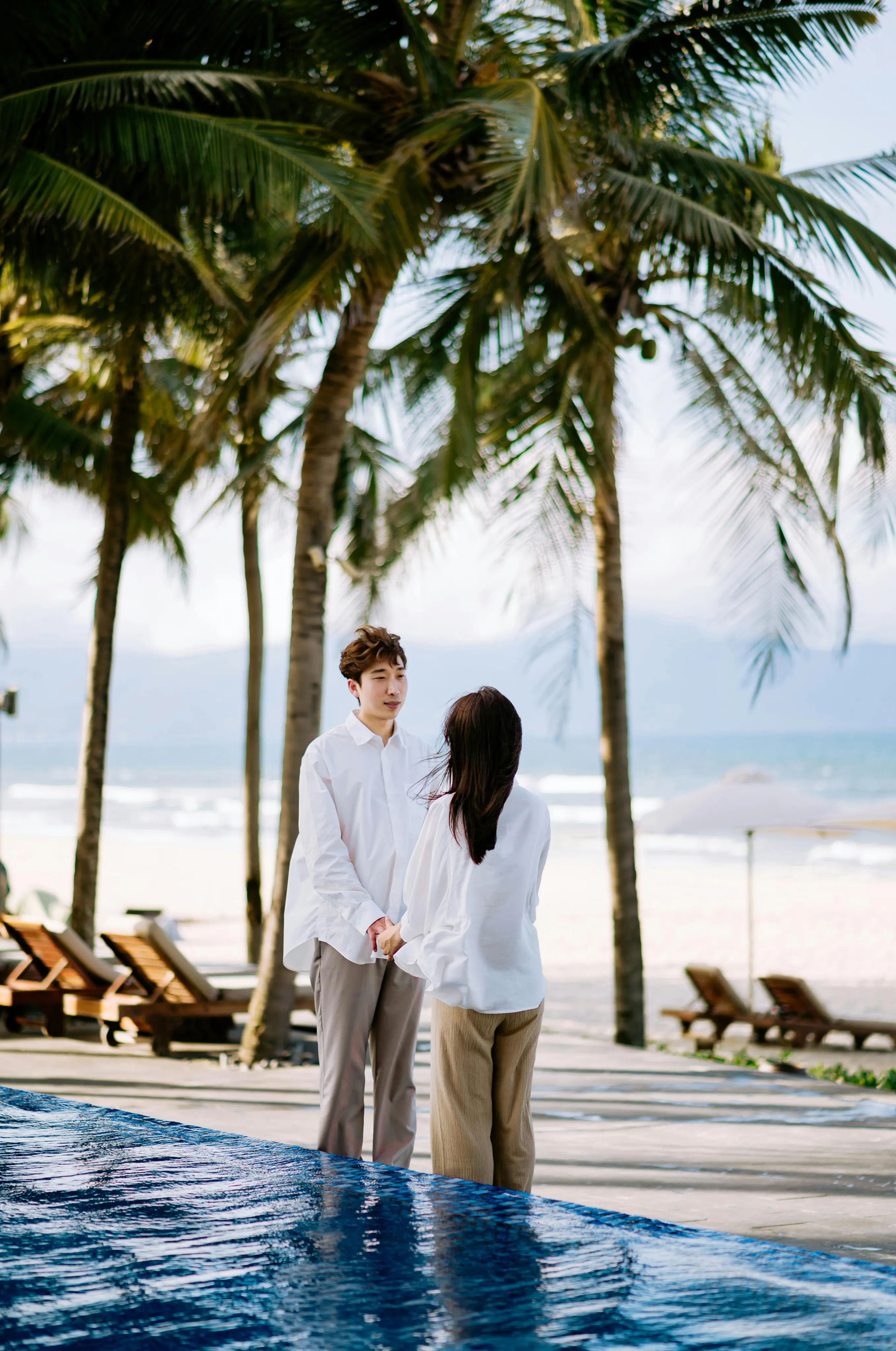Couple holding hands near a pool with palm trees and a beach in the background.