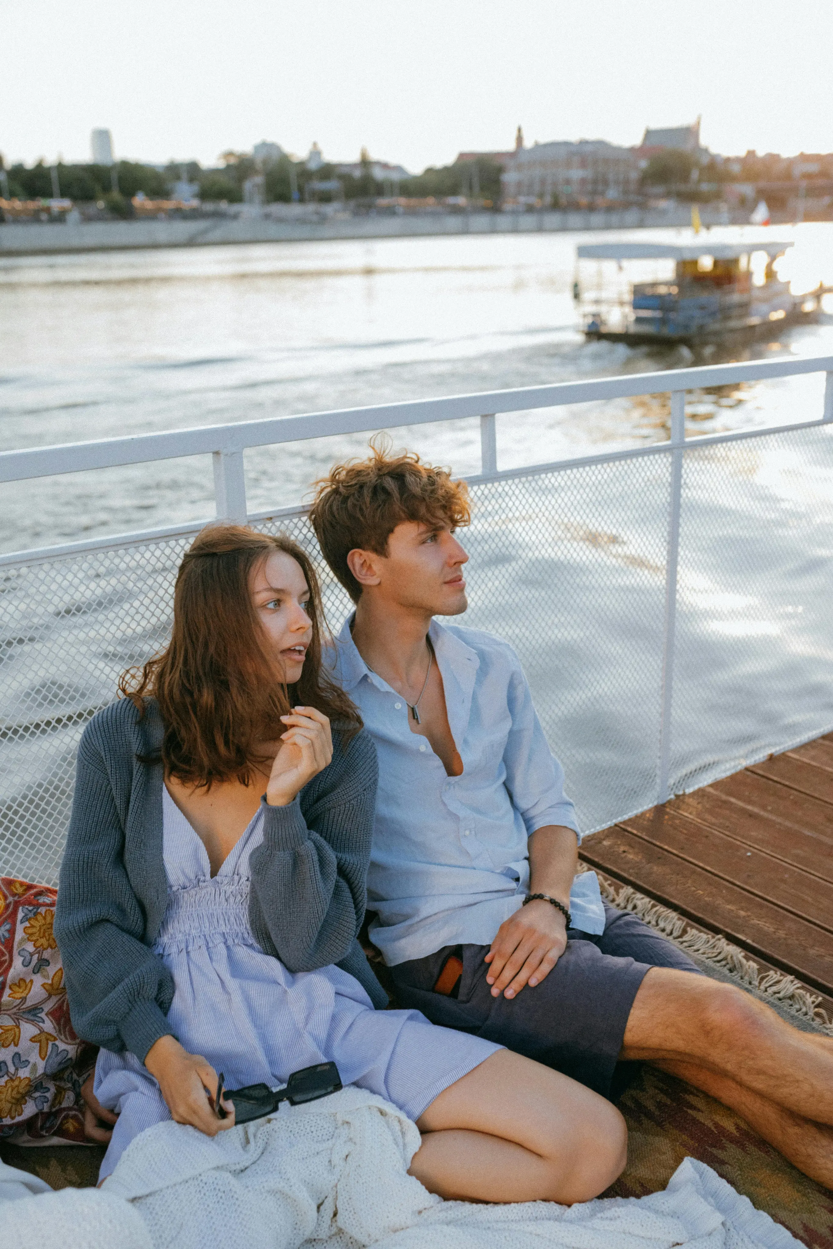 Young couple sitting on a blanket by the waterfront, looking to the side during sunset.