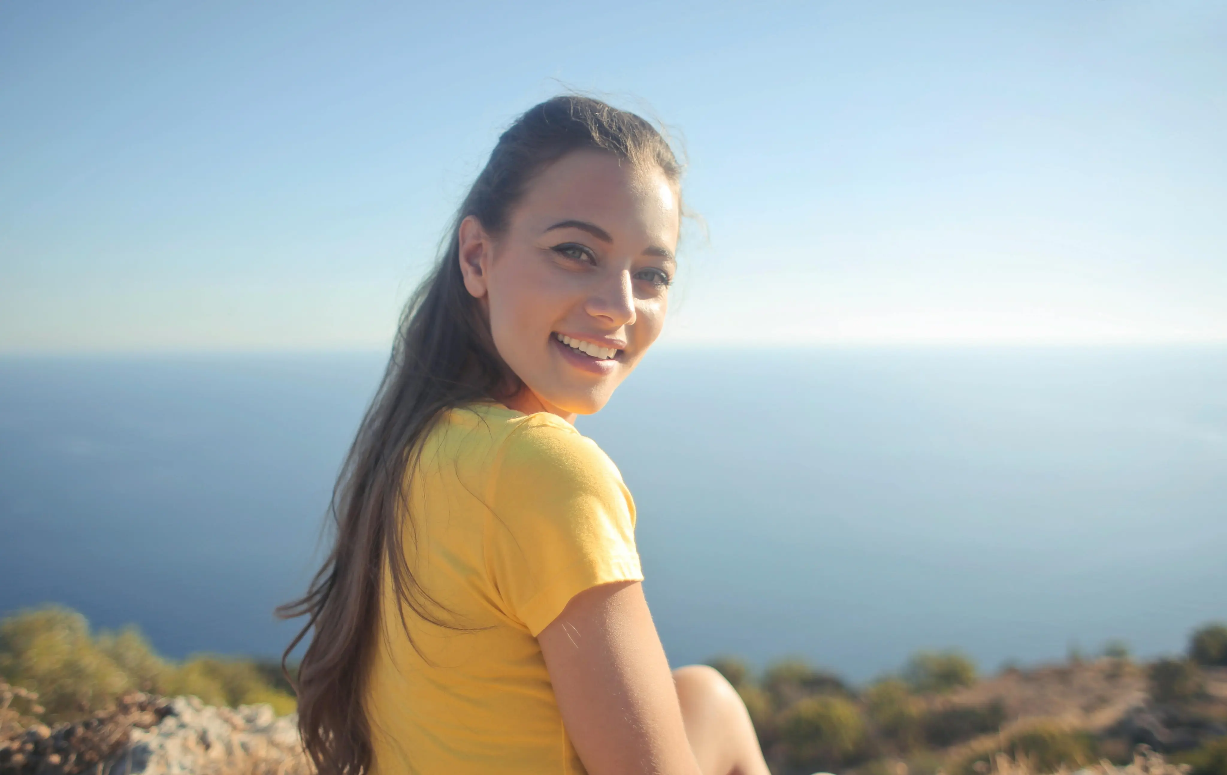 Smiling young woman in a yellow shirt sitting outdoors with a sea and clear sky in the background.