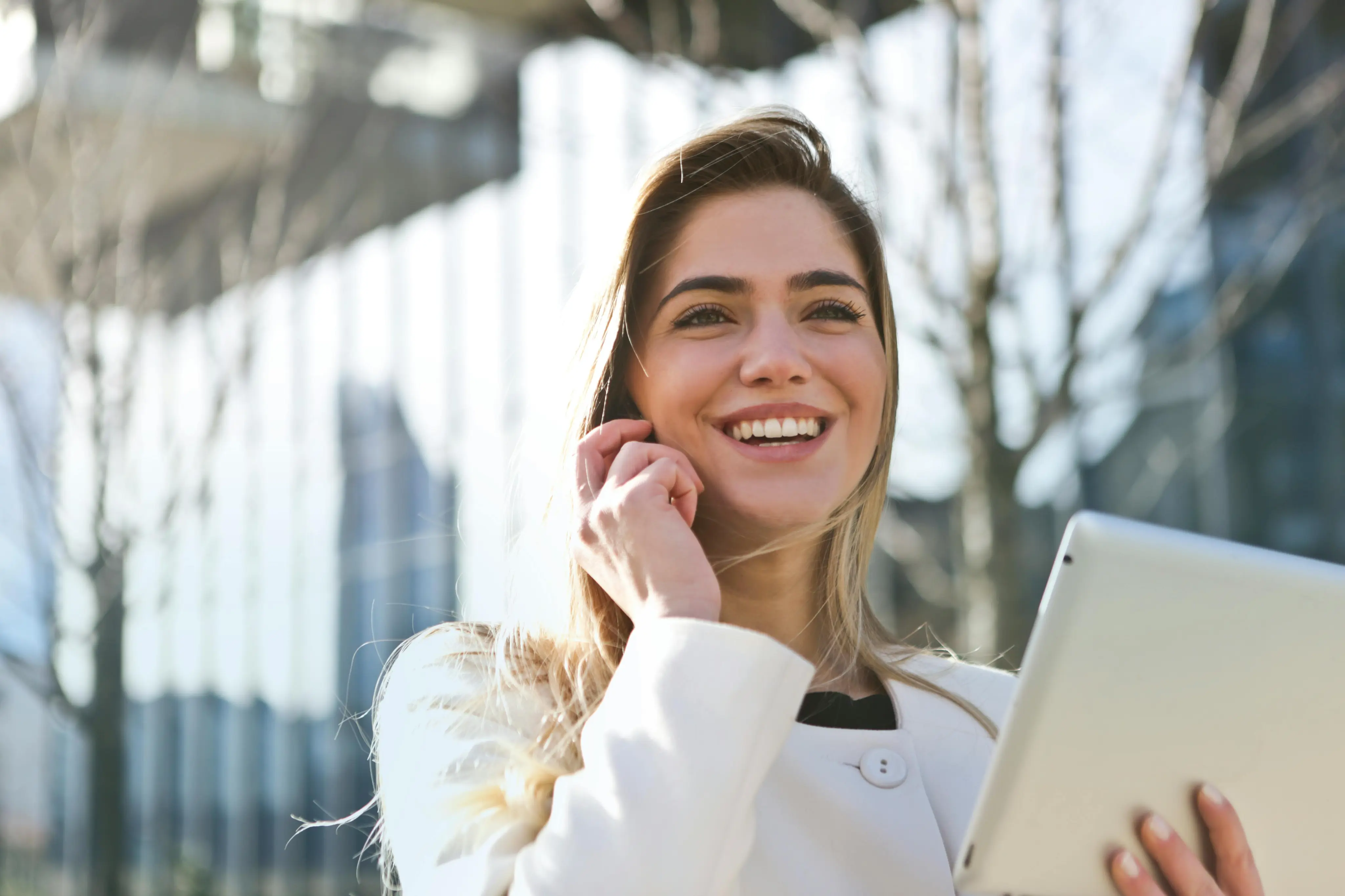 Smiling woman holding a tablet and touching her face outdoors in a bright urban setting.