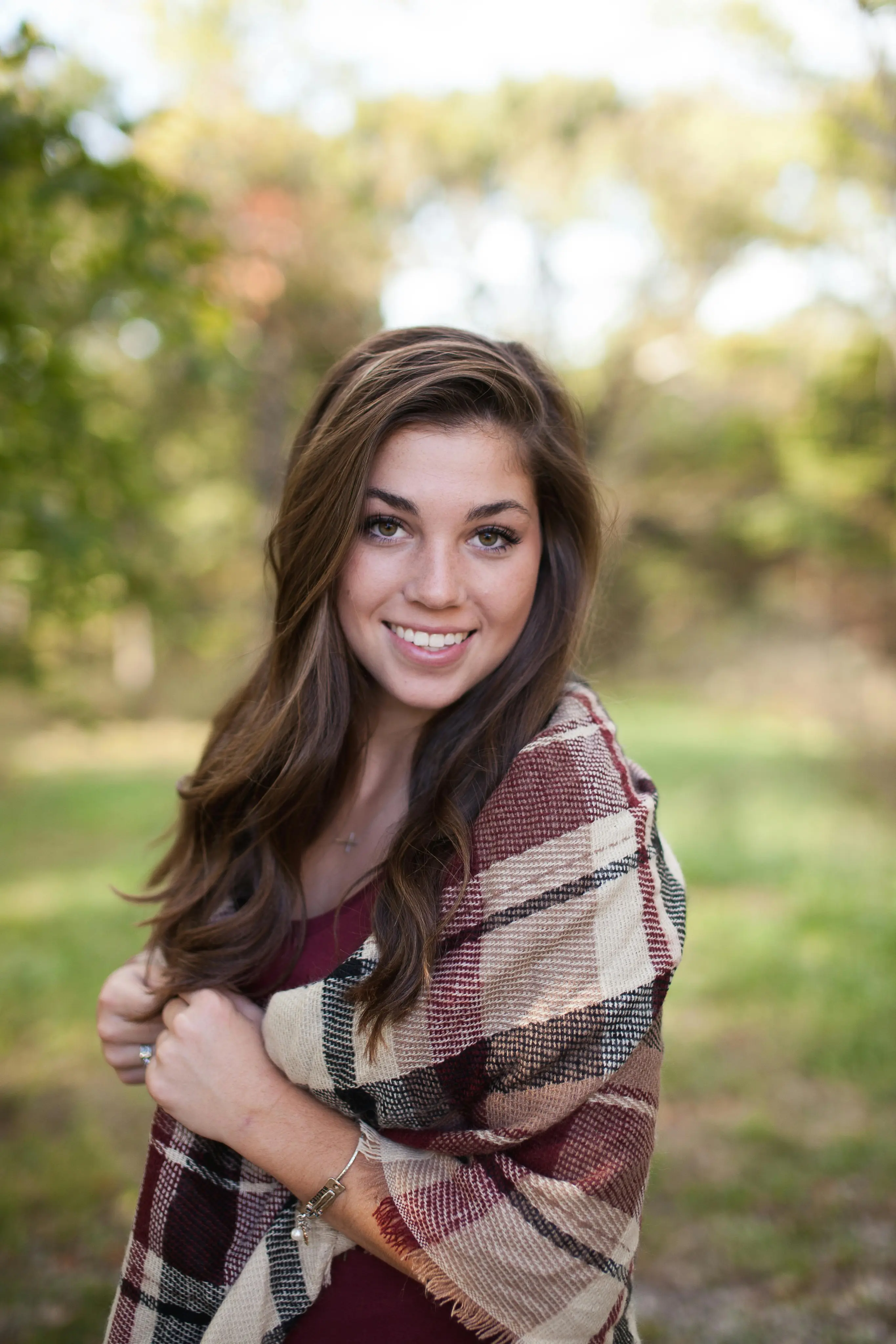 Smiling young woman with long brown hair wrapped in a plaid shawl outdoors with blurred greenery background.