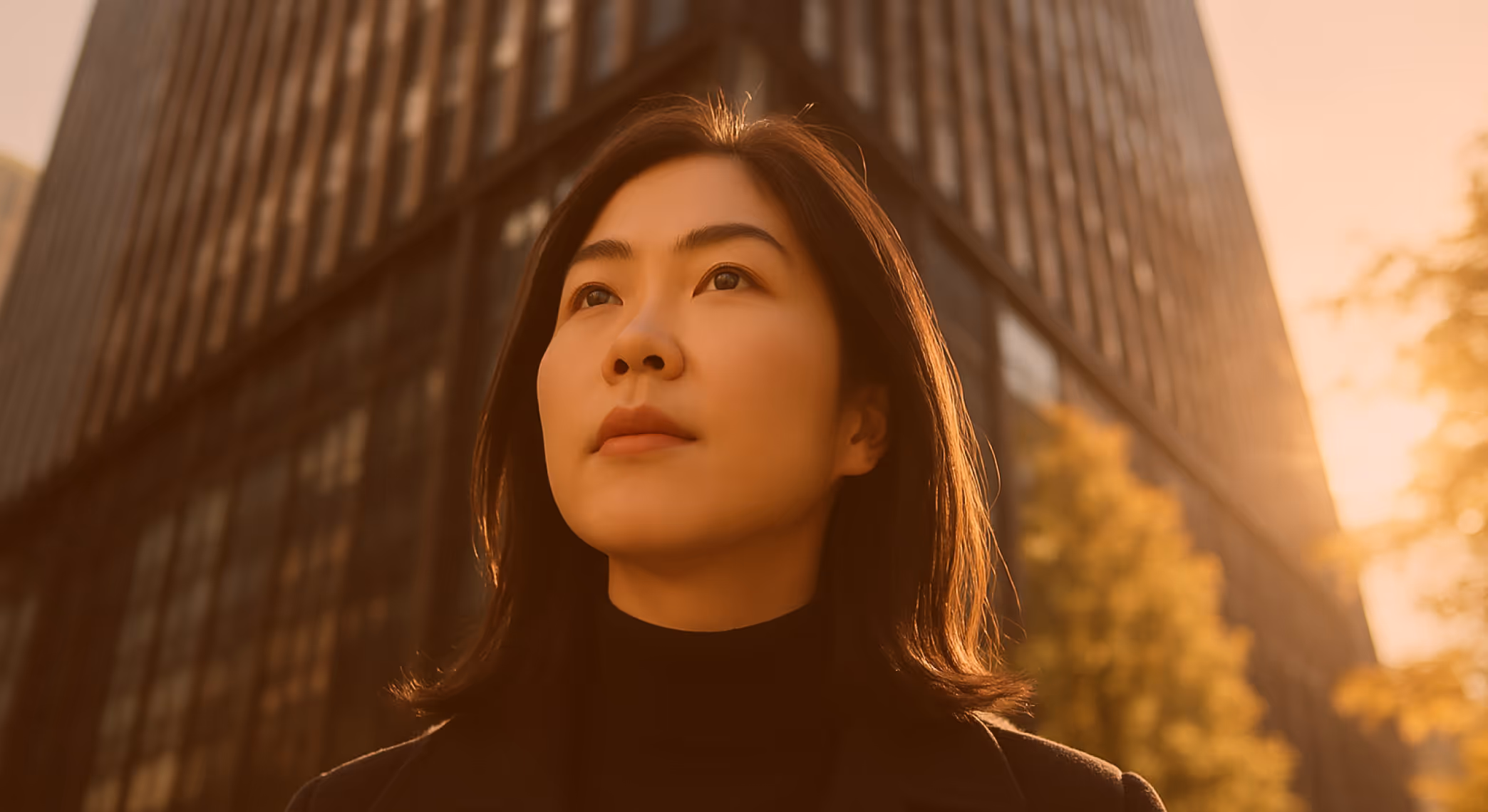 Young woman with dark hair looking thoughtfully upward with a blurred tall building and trees in the background at sunset.