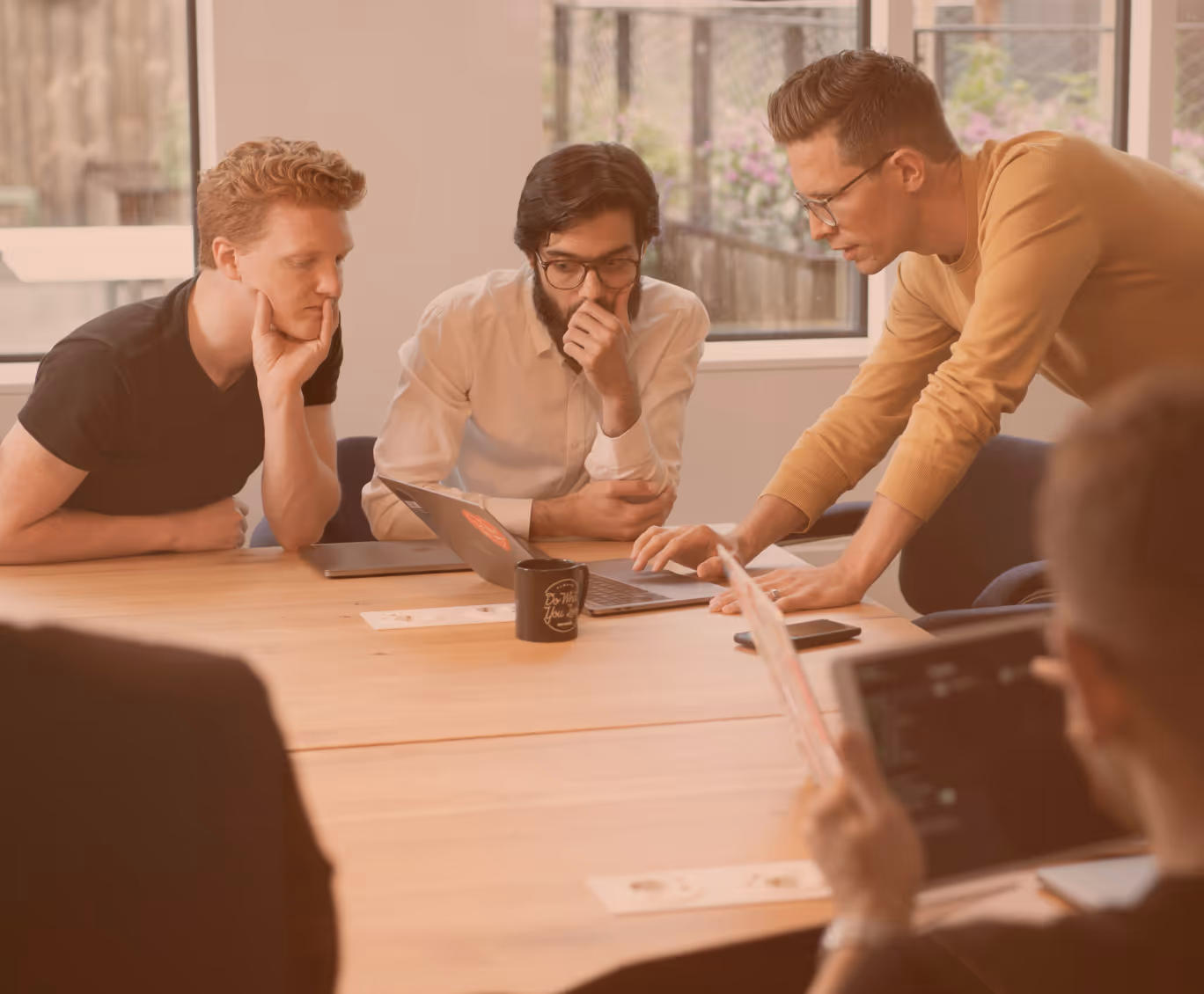 Three men intensely discussing and looking at a laptop on a wooden table in a bright office room.