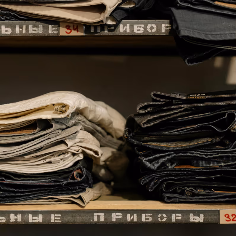 Stacks of folded pants in various shades, including beige and dark denim, arranged on wooden shelves.