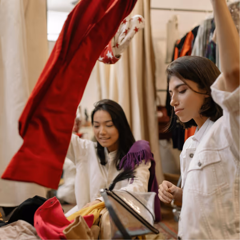 Two women browsing through clothes in a store, holding up garments to examine.
