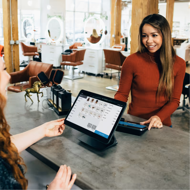 A smiling woman in a red sweater stands behind a salon counter with a tablet showing a service menu to a seated customer.