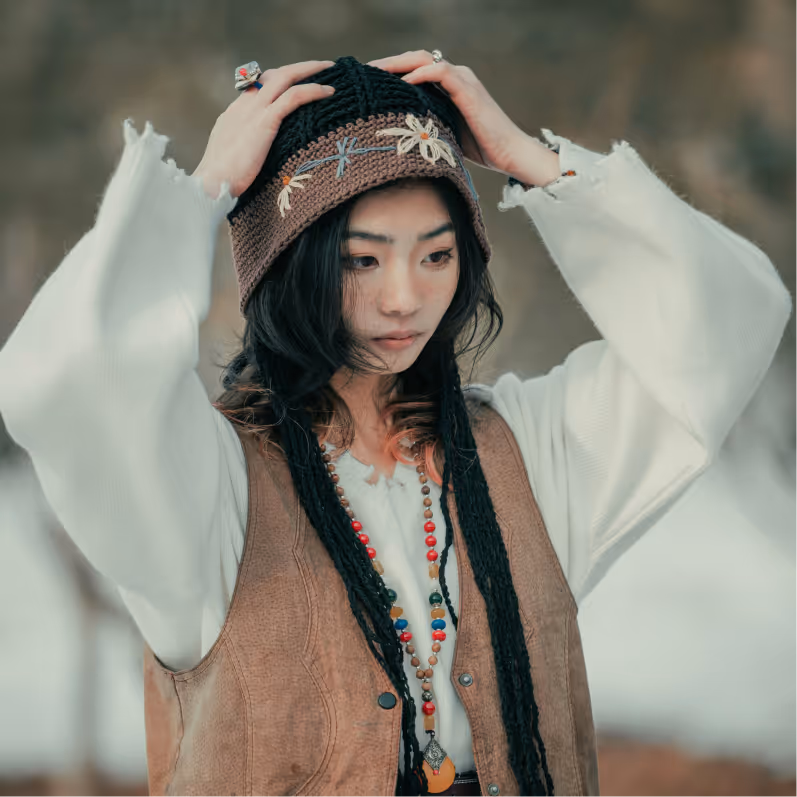 Young woman wearing a white blouse, brown vest, bead necklaces, and a knitted hat with floral embroidery, adjusting the hat with both hands.