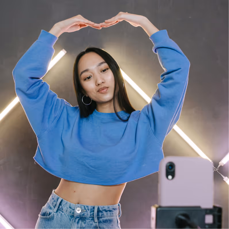 Young woman in blue cropped sweatshirt and jeans making a heart shape with her hands above her head while taking a selfie with a smartphone on a tripod.