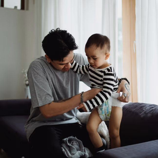 Young adult helping toddler in striped shirt and diaper to stand on a sofa in a living room.