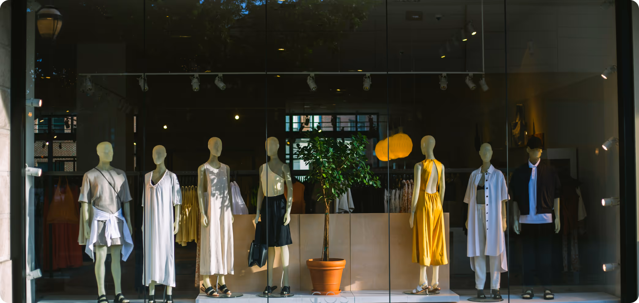 Store window display featuring six mannequins dressed in summer outfits including dresses, skirts, and casual tops with a potted plant in the center.